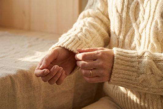 Close-up of hands adjusting the cuff of a cream cable-knit sweater in warm window light