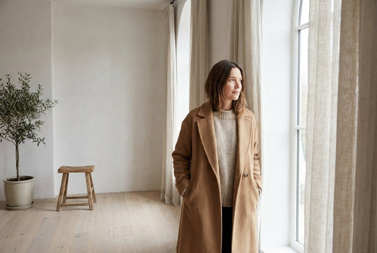 Woman in a camel wool coat over an oatmeal knit standing beside a tall window